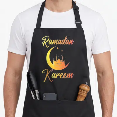 Kareem eid al fitr apron - Yellow orange crescent moon with mosque silhouette and Ramadan Kareem text — shown on a black cotton canvas apron against a white background