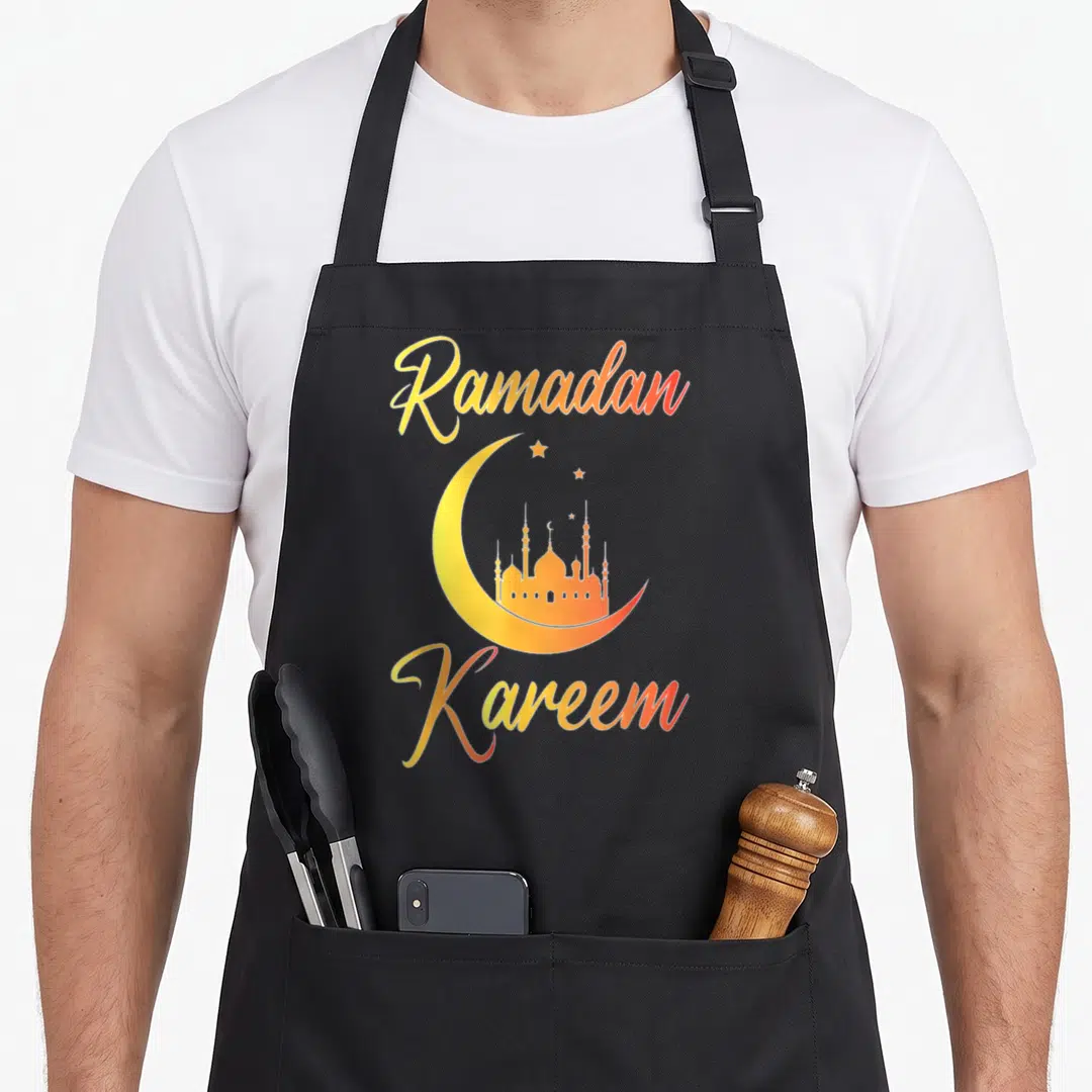 Kareem eid al fitr apron - Yellow orange crescent moon with mosque silhouette and Ramadan Kareem text — shown on a black cotton canvas apron against a white background