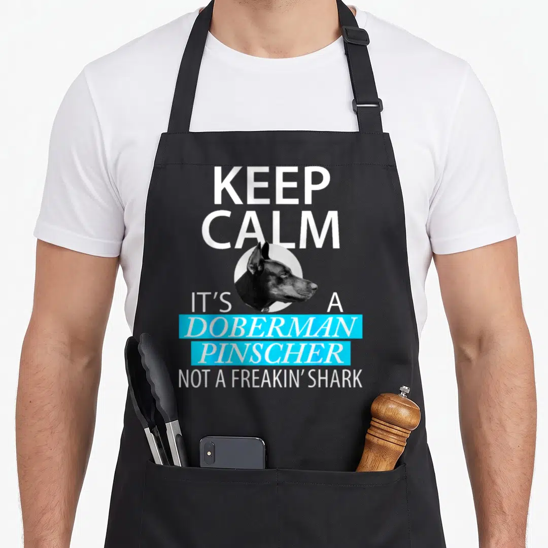 Funny doberman pinscher apron - Doberman head in circle with white and cyan stacked text reading Keep Calm It's — shown on a black cotton canvas apron against a white background