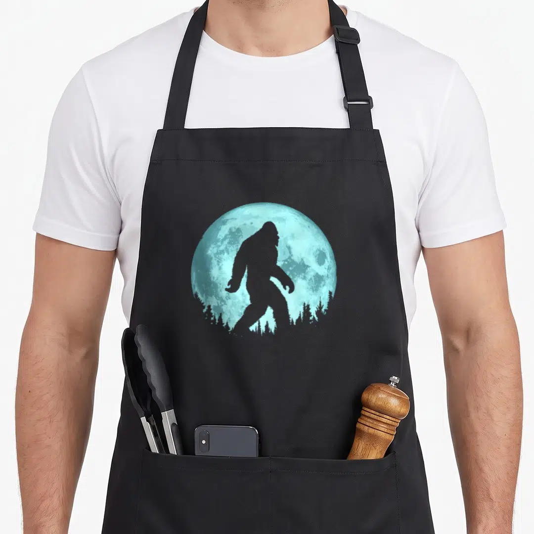 Minimalist tree apron - Grey Bigfoot silhouette walking past light blue full moon and pine tree line — shown on a black cotton canvas apron against a white background
