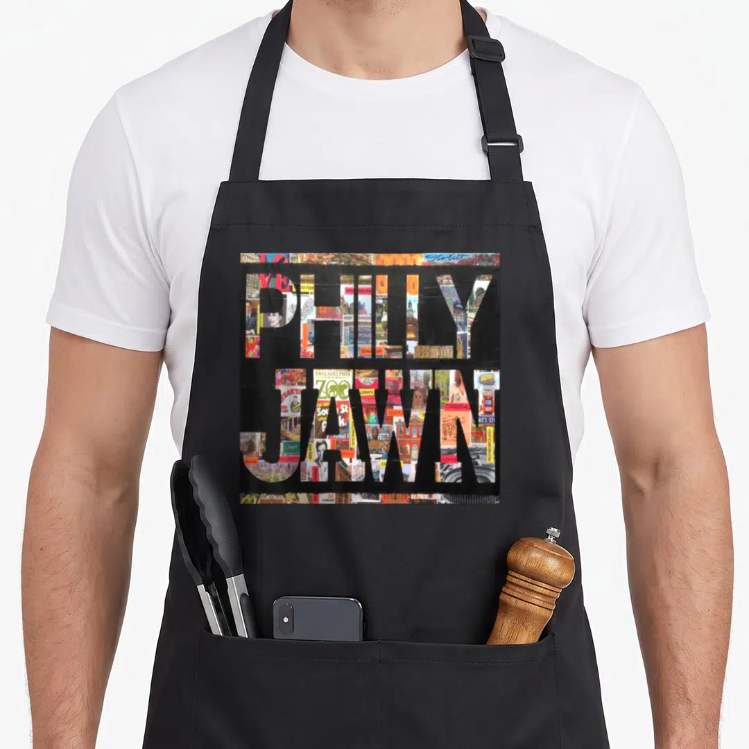 Jawn phillies apron - Stacked PHILLY and JAWN text filled with city photo collage in black, red, blue, yellow, and white — shown on a black cotton canvas apron against a white background