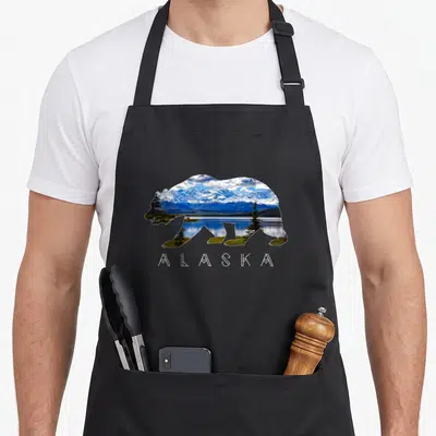 Alaskan bear apron - Bear silhouette filled with blue mountains and green trees above white text — shown on a black cotton canvas apron against a white background
