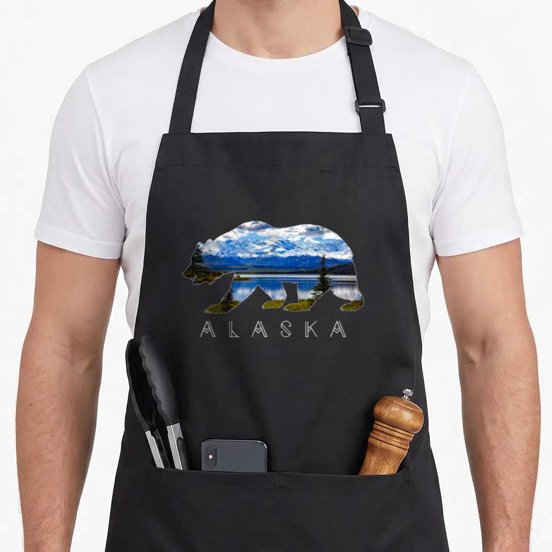 Alaskan bear apron - Bear silhouette filled with blue mountains and green trees above white text — shown on a black cotton canvas apron against a white background