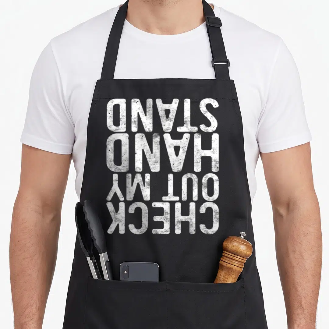 Handstand gymnastics apron - White distressed text stacked upside down reading CHECK OUT MY HANDSTAND — shown on a black cotton canvas apron against a white background