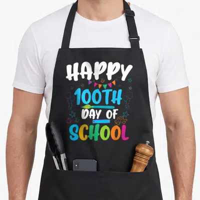 Vintage 100th teacher apron - HAPPY 100TH DAY OF SCHOOL text with bunting, ladybug, pencil, ruler, car, and ball — shown on a black cotton canvas apron against a white background