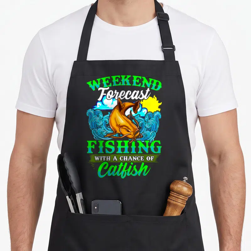 Catfish fishing aprons - Cartoon orange catfish jumping from blue waves under green and blue stacked text — shown on a black cotton canvas apron against a white background