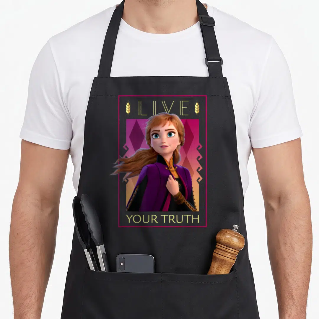 Truth frozen apron - Anna in purple cape framed by pink border with Live Your Truth text and gold wheat stalks — shown on a black cotton canvas apron against a white background