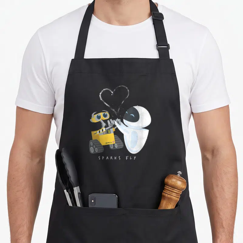 Fly e.a apron - WALL-E and EVE facing each other with dotted heart and SPARKS FLY text — shown on a black cotton canvas apron against a white background
