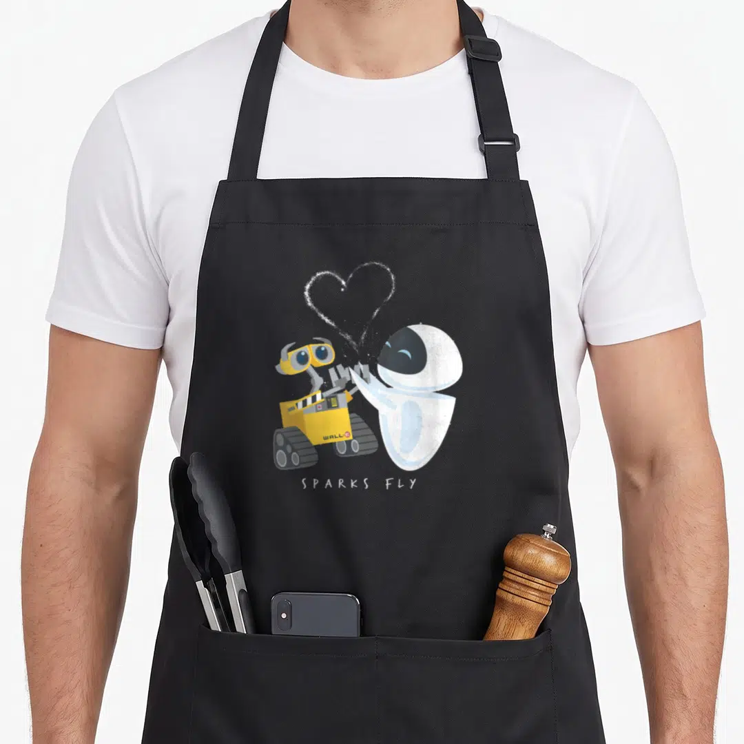 Fly e.a apron - WALL-E and EVE facing each other with dotted heart and SPARKS FLY text — shown on a black cotton canvas apron against a white background