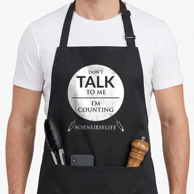 Operating nurse apron - DON'T TALK TO ME I'M COUNTING text in white circle with two syringes and hashtag — shown on a black cotton canvas apron against a white background