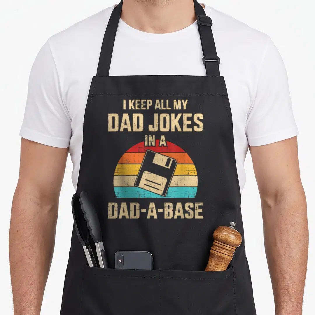 Base joke apron - Beige floppy disk over red orange yellow blue sunset with dad joke pun text — shown on a black cotton canvas apron against a white background