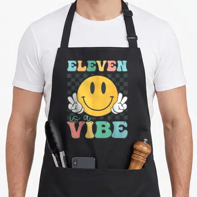 Eleven custom face apron - Yellow smiley face with peace sign hands and colorful ELEVEN is a VIBE text on grey — shown on a black cotton canvas apron against a white background