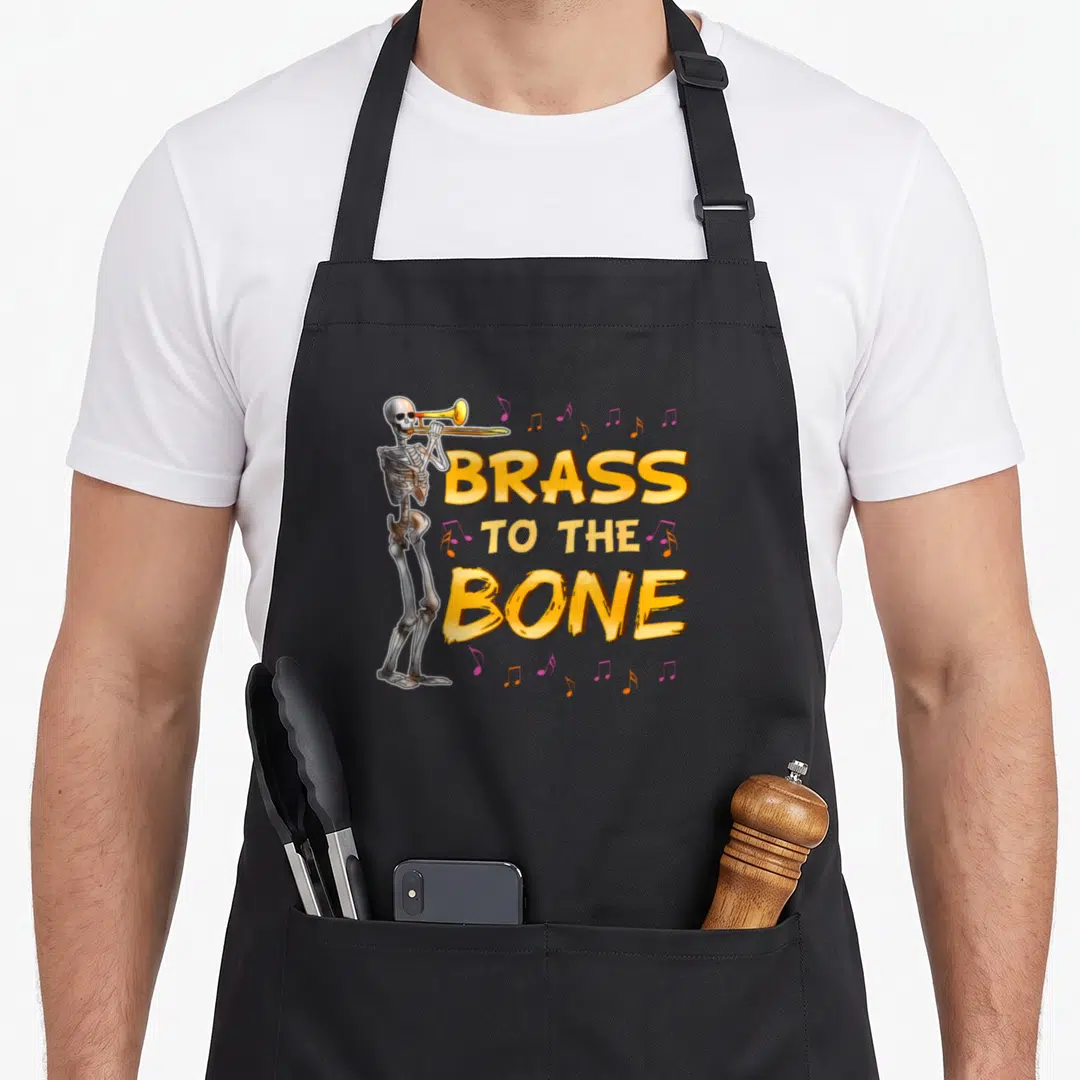 Trombone skeleton apron - Cartoon skeleton playing trumpet next to yellow text BRASS TO THE BONE and pink notes — shown on a black cotton canvas apron against a white background