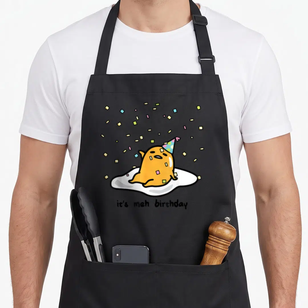 Gudetama egg apron - Orange blob creature in party hat on fried egg with it's meh birthday text and confetti — shown on a black cotton canvas apron against a white background