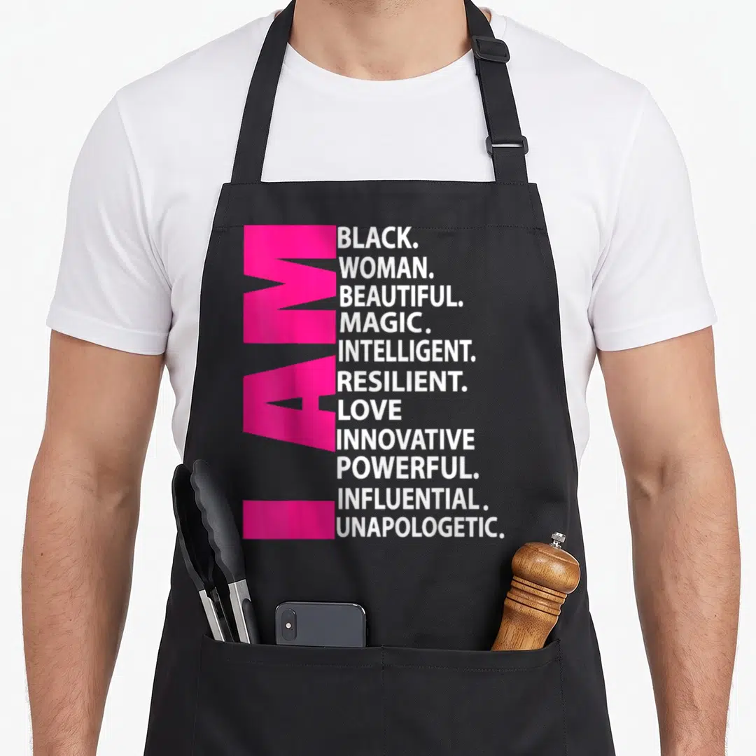 Afro magic apron - Vertical pink I AM text next to stacked white adjectives including BLACK WOMAN BEAUTIFUL MAGIC — shown on a black cotton canvas apron against a white background