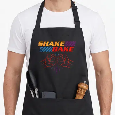Shake friends apron - SHAKE AND BAKE text with red fists and checkered flags in orange blue and purple — shown on a black cotton canvas apron against a white background