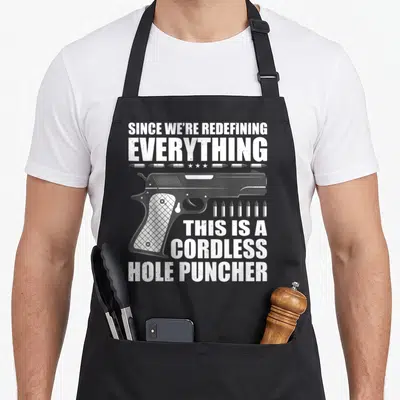 Redefining hole apron - SINCE WE'RE REDEFINING EVERYTHING CORDLESS HOLE PUNCHER text with handgun and bullets — shown on a black cotton canvas apron against a white background