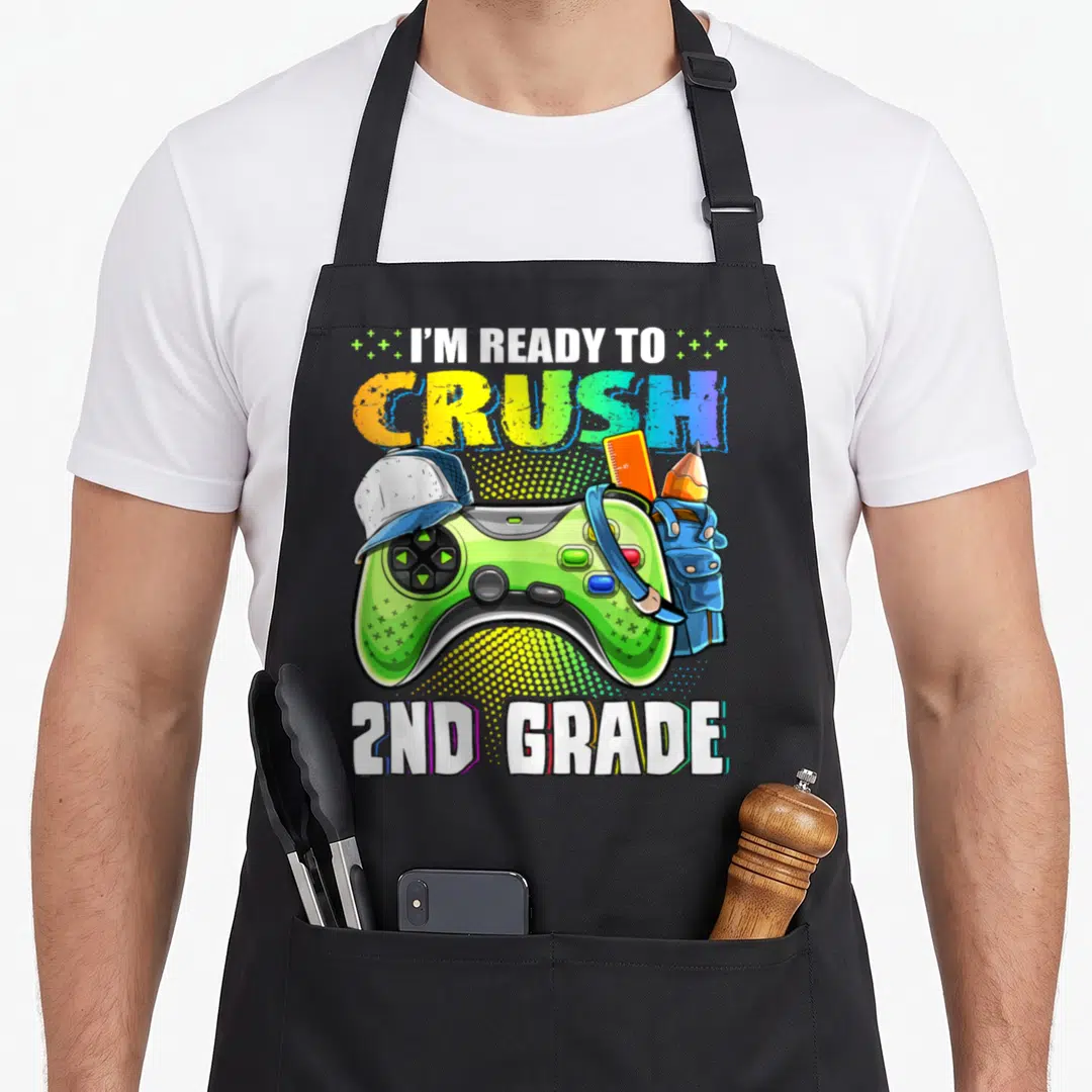 Crush 2nd video game apron - Green controller with cap, ruler, pencil, backpack, and I'M READY TO CRUSH 2ND GRADE text — shown on a black cotton canvas apron against a white background