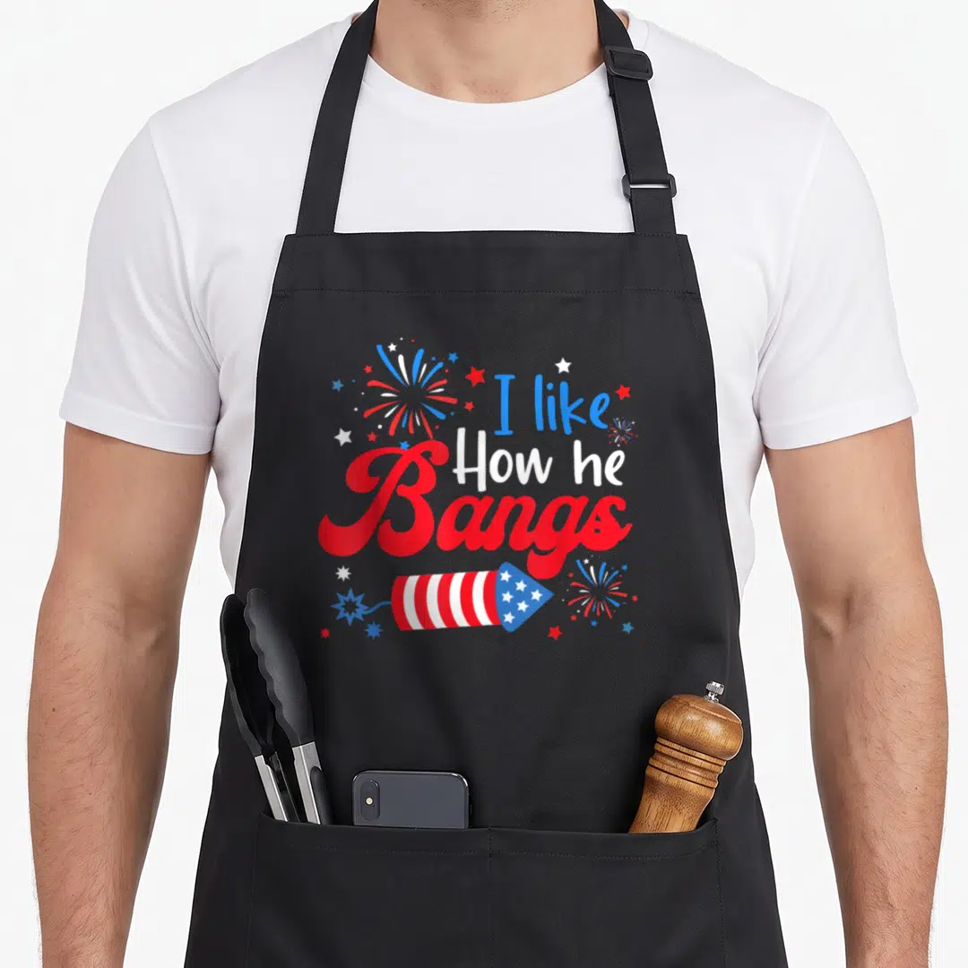 Explode 4th of july apron - I like How he Bangs text with firework rocket and stars in red white blue — shown on a black cotton canvas apron against a white background