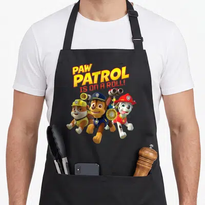 Roll novelty apron - Stacked PAW PATROL IS ON A ROLL text above three running cartoon puppies in colored uniforms — shown on a black cotton canvas apron against a white background