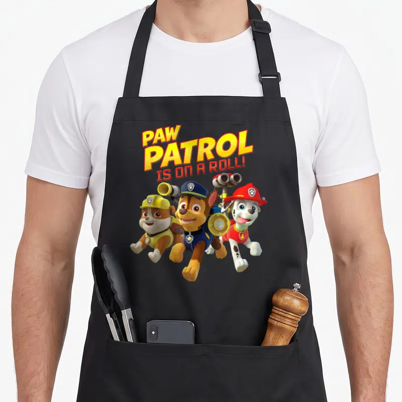 Roll novelty apron - Stacked PAW PATROL IS ON A ROLL text above three running cartoon puppies in colored uniforms — shown on a black cotton canvas apron against a white background