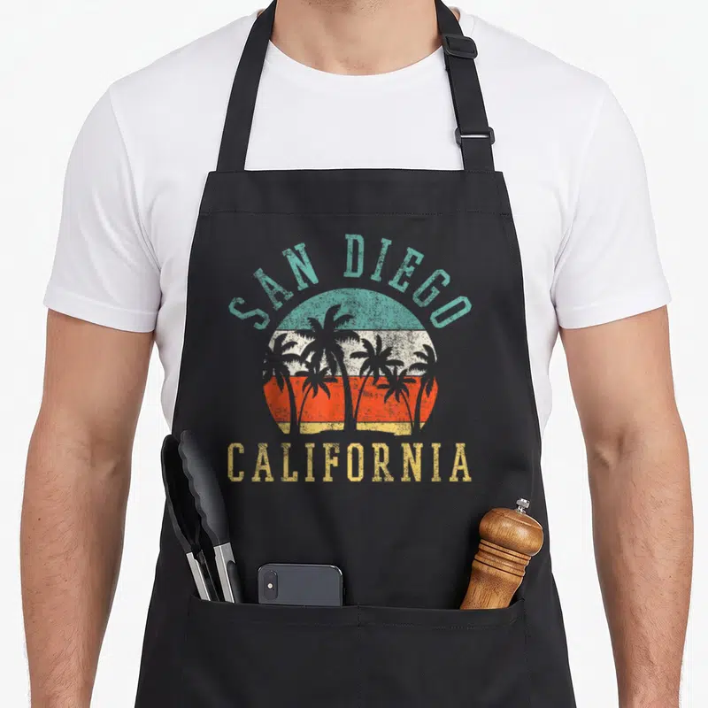 Funny california san diego apron - Retro sunset with five palm trees and arching SAN DIEGO CALIFORNIA text — shown on a black cotton canvas apron against a white background