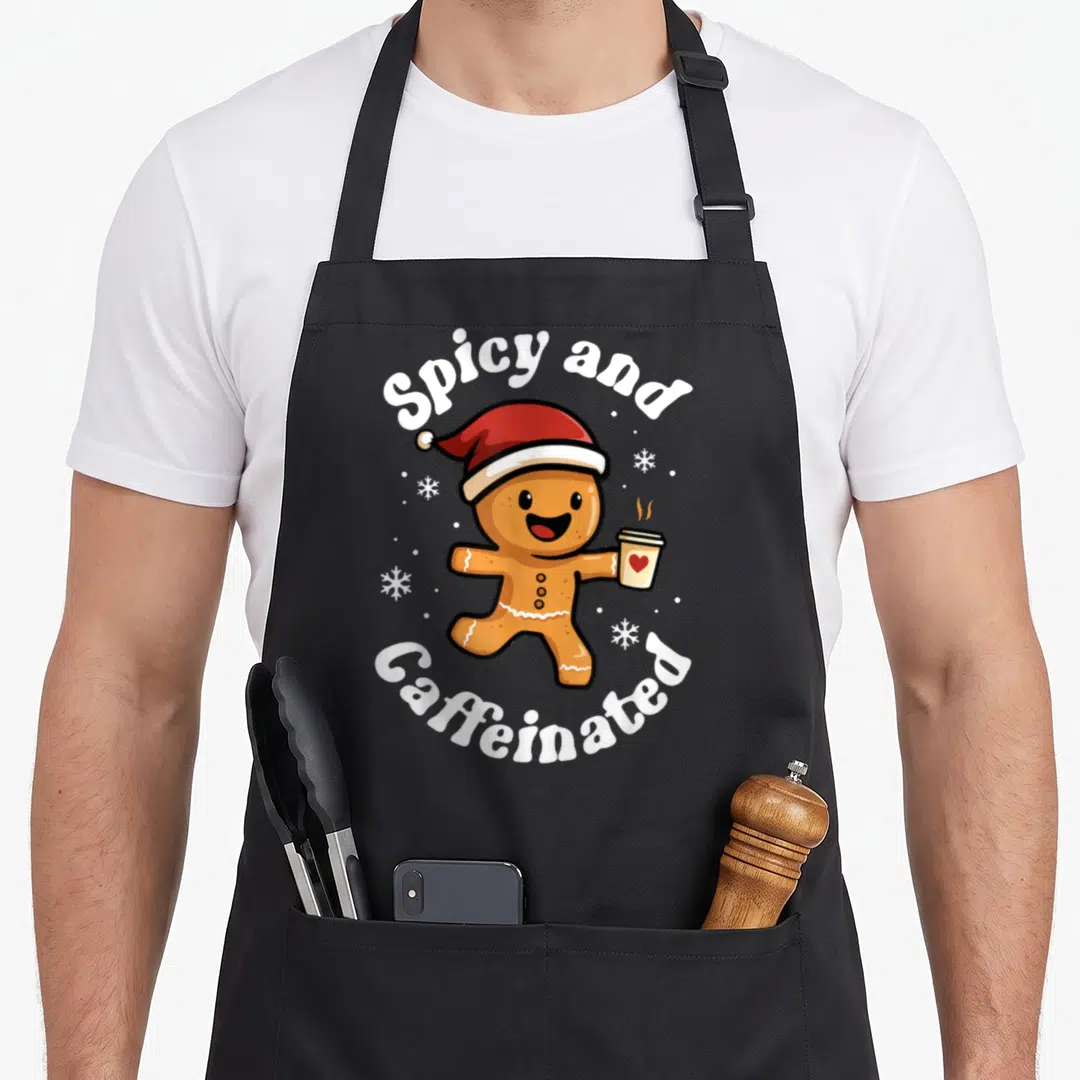 Spicy gingerbread apron - Cartoon gingerbread man in red hat holding coffee cup with arched Spicy and Caffeinated text — shown on a black cotton canvas apron against a white background