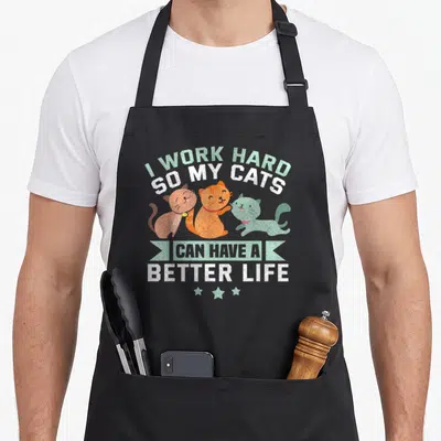 Hard better cat apron - Three cartoon cats in brown orange and mint below stacked text and ribbon banner — shown on a black cotton canvas apron against a white background
