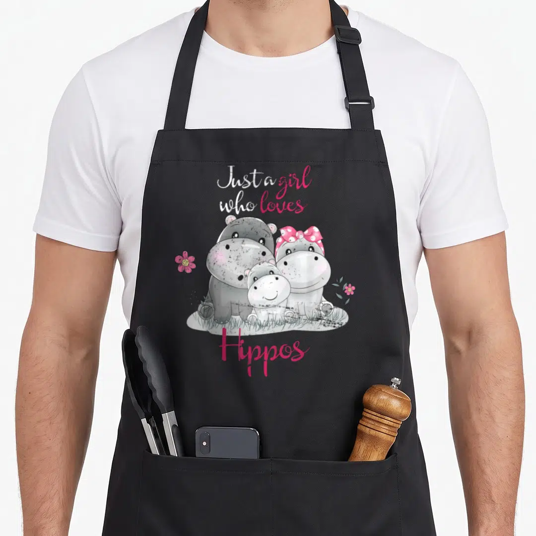 Cute hippo apron - Three cartoon hippos standing in grass with pink flowers and text Just a girl who loves Hippos — shown on a black cotton canvas apron against a white background