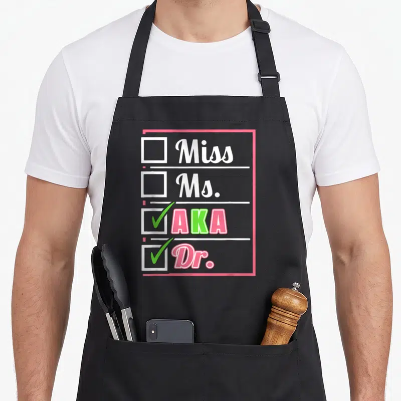 Aka doctor apron - Checklist with checked boxes for AKA and Dr. in pink and green text — shown on a black cotton canvas apron against a white background