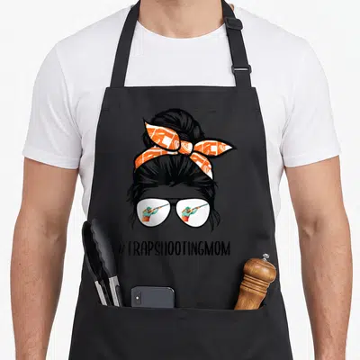 Trap messy aprons - Graphic of messy bun, patterned bandana, glasses reflecting shooter, and #TRAPSHOOTINGMOM text — shown on a black cotton canvas apron against a white background