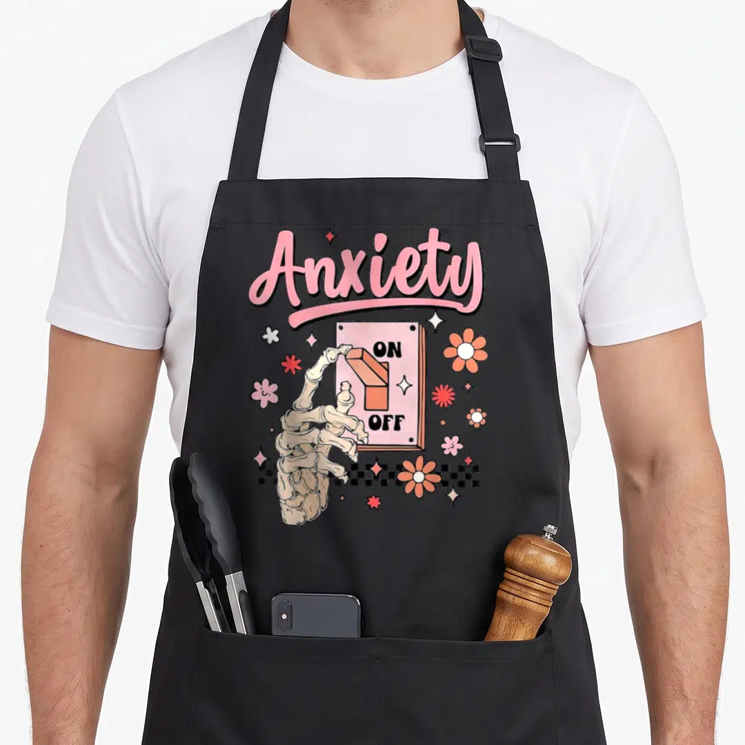 Switch anxiety awareness apron - Skeleton hand flipping ON OFF switch with pink Anxiety script and scattered flowers — shown on a black cotton canvas apron against a white background