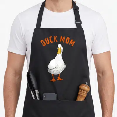 Illustration duck apron - Arched orange DUCK MOM text above a standing white cartoon duck with orange feet — shown on a black cotton canvas apron against a white background