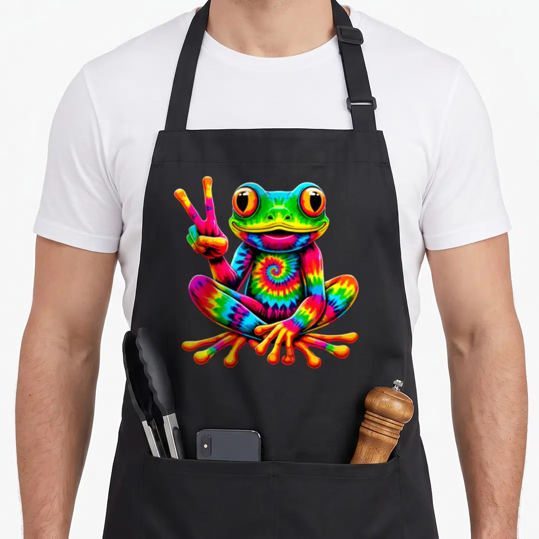 Frog tie-dye apron - Rainbow tie-dye frog sitting and holding up a peace sign in cartoon style — shown on a black cotton canvas apron against a white background