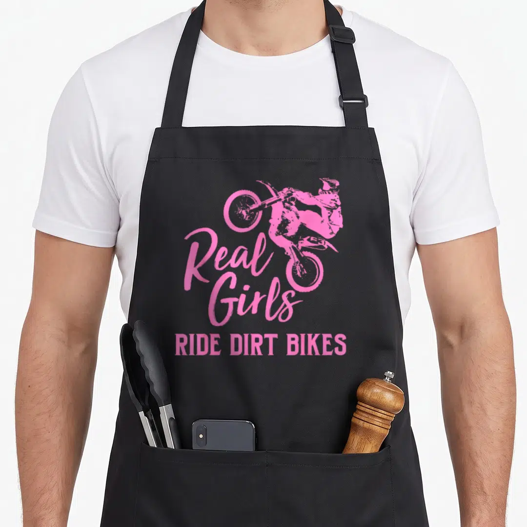 Funny ride bike apron - Pink cartoon dirt bike rider jumping above Real Girls RIDE DIRT BIKES text — shown on a black cotton canvas apron against a white background