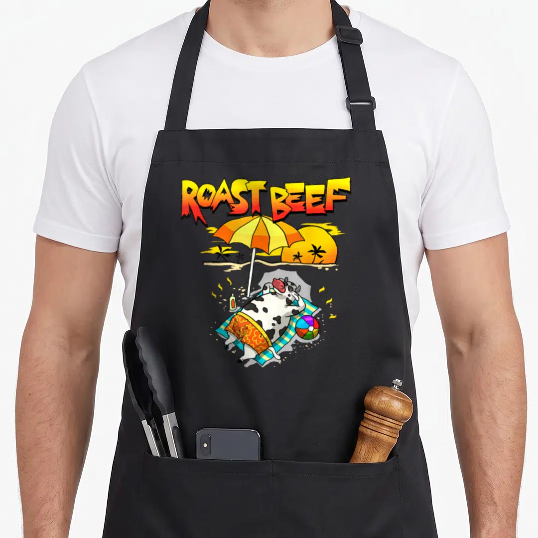 Roast cow apron - Cartoon cow sunbathing on beach towel under umbrella with ROAST BEEF text — shown on a black cotton canvas apron against a white background