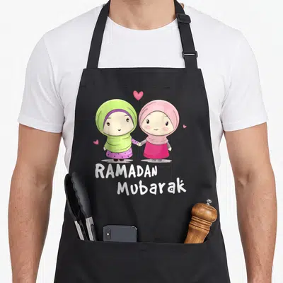 Ramadan cartoon apron - Two cartoon girls in hijabs holding hands with RAMADAN Mubarak text and hearts — shown on a black cotton canvas apron against a white background