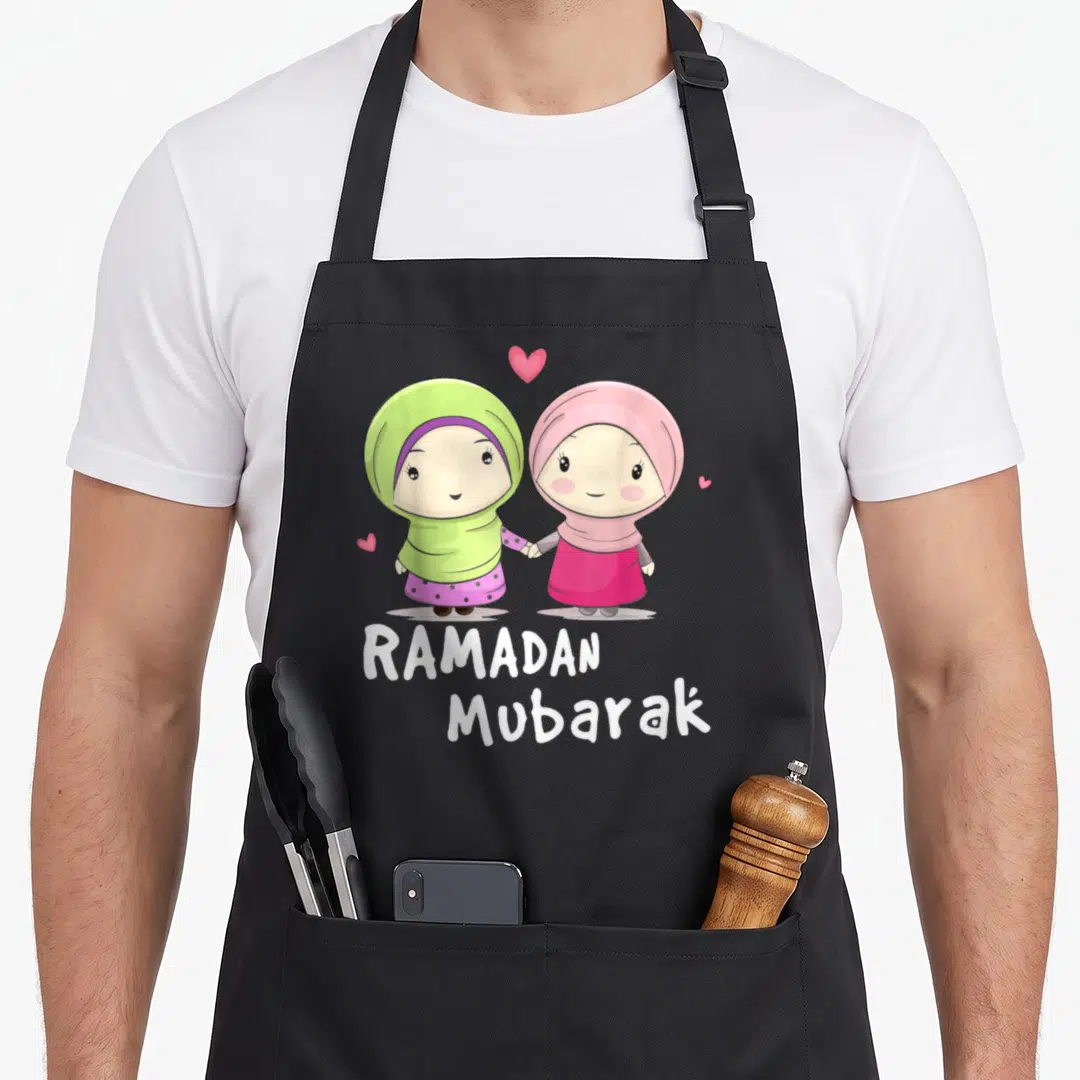 Ramadan cartoon apron - Two cartoon girls in hijabs holding hands with RAMADAN Mubarak text and hearts — shown on a black cotton canvas apron against a white background