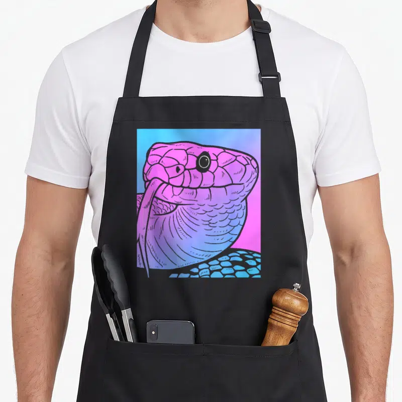 Snake goth apron - Cartoon snake head in pink and purple scales against a blue and pink gradient background — shown on a black cotton canvas apron against a white background