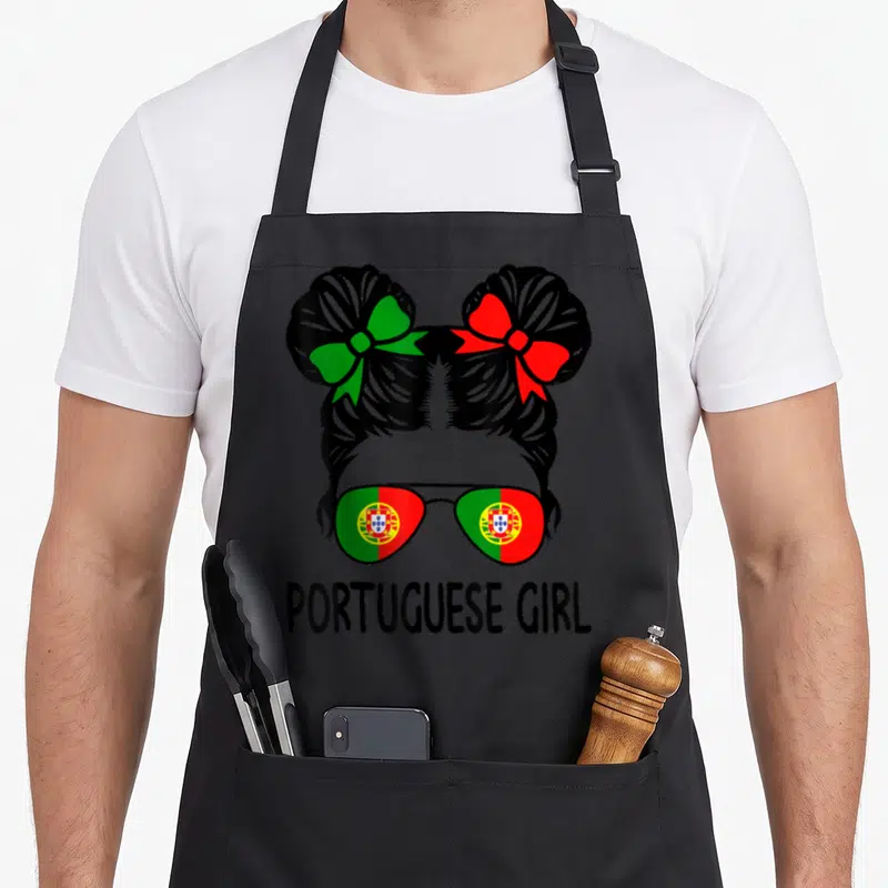 Messy portuguese apron - Cartoon girl with two buns, green and red bows, flag sunglasses, and PORTUGUESE GIRL text — shown on a black cotton canvas apron against a white background