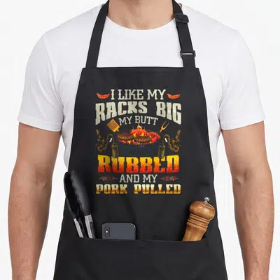 Rubbed i love my pig apron - Stacked grilling pun text surrounding a charcoal grill with spatula and tongs in red — shown on a black cotton canvas apron against a white background