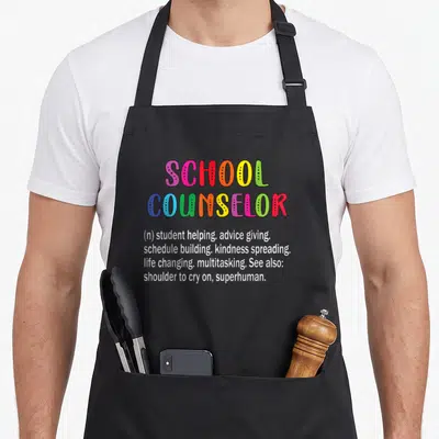 Definition school counselor apron - Rainbow text reading School Counselor with definition list including kindness — shown on a black cotton canvas apron against a white background