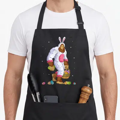 Bigfoot egg apron - Cartoon bigfoot in white bunny suit holding basket and eggs with stars — shown on a black cotton canvas apron against a white background