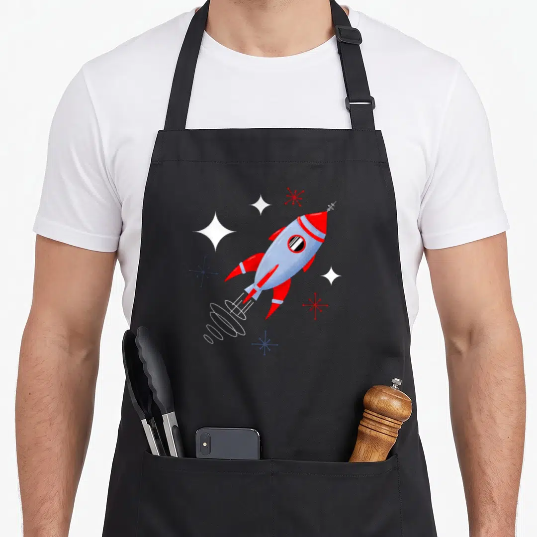 Rocket modern apron - Red and blue retro rocket with white stars and atomic sparkles on stiff canvas — shown on a black cotton canvas apron against a white background