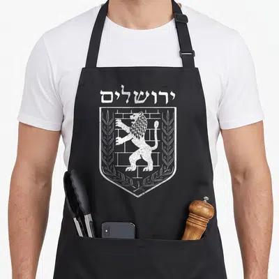 Emblem jewish apron - White distressed shield with standing lion, brick wall, olive branches, and Hebrew text on black — shown on a black cotton canvas apron against a white background