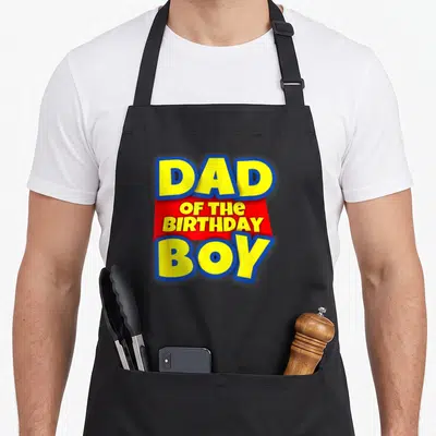 Birthday apron with pockets for women - Yellow DAD BOY text with blue outlines and red OF THE BIRTHDAY banner — shown on a black cotton canvas apron against a white background