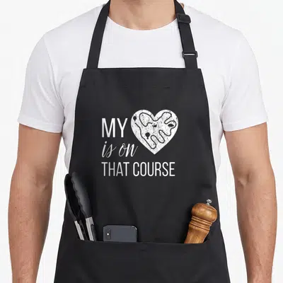 Course country apron - White heart with winding track path beside text MY is on THAT COURSE — shown on a black cotton canvas apron against a white background