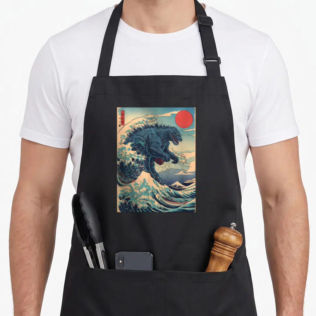 Kaiju wave apron - Godzilla emerging from a blue wave with a red sun and mountain background — shown on a black cotton canvas apron against a white background