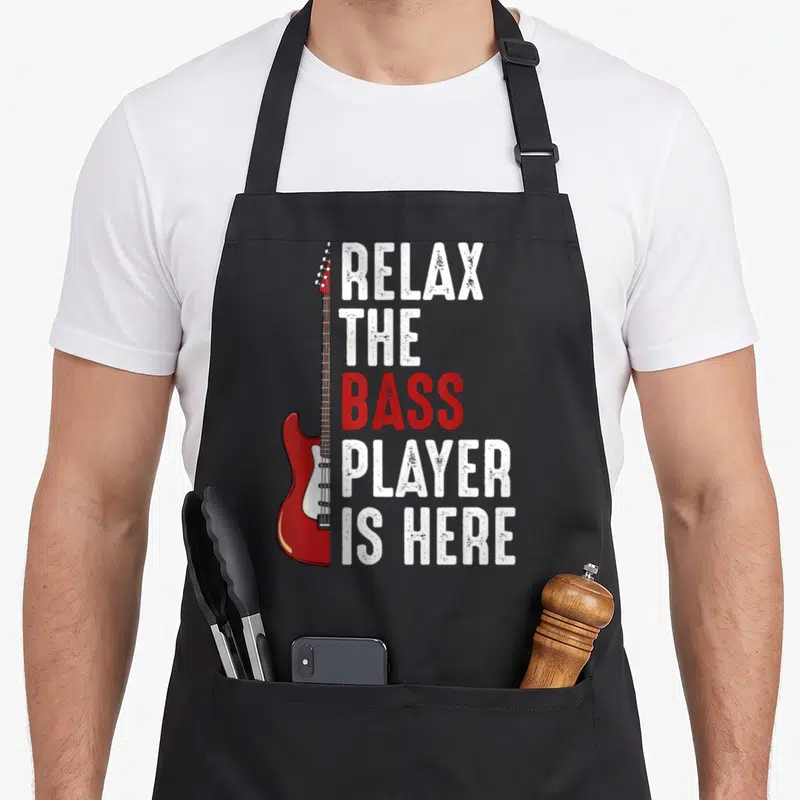 Relax bass apron - Red electric bass guitar next to stacked white and red distressed text — shown on a black cotton canvas apron against a white background