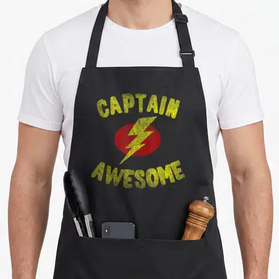 Captain flippin sweet gear apron - Retro yellow Captain Awesome text arching over red oval with yellow lightning bolt — shown on a black cotton canvas apron against a white background